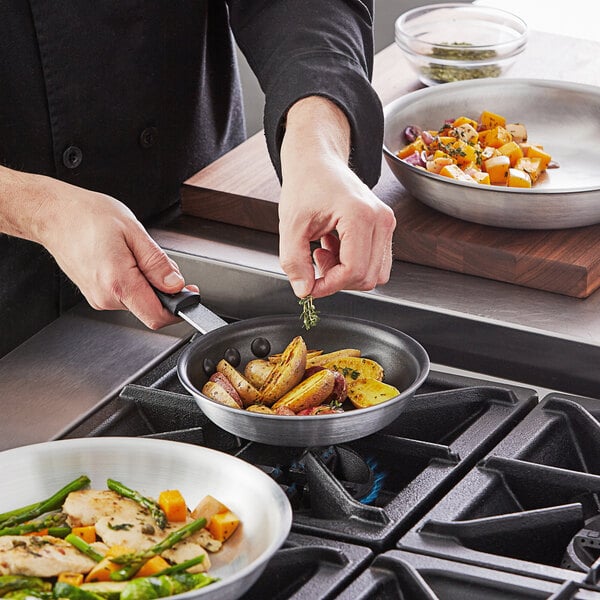 A person cooking food in a Vollrath Arkadia aluminum non-stick fry pan with a black silicone handle.