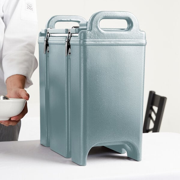 A person holding a bowl of food next to a blue Cambro insulated soup carrier.