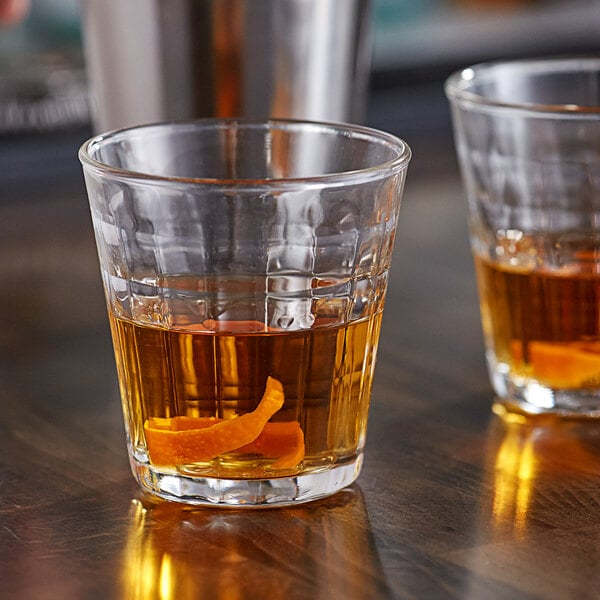 Two Duralex stackable glass tumblers filled with liquid and garnished with orange peel on a table.