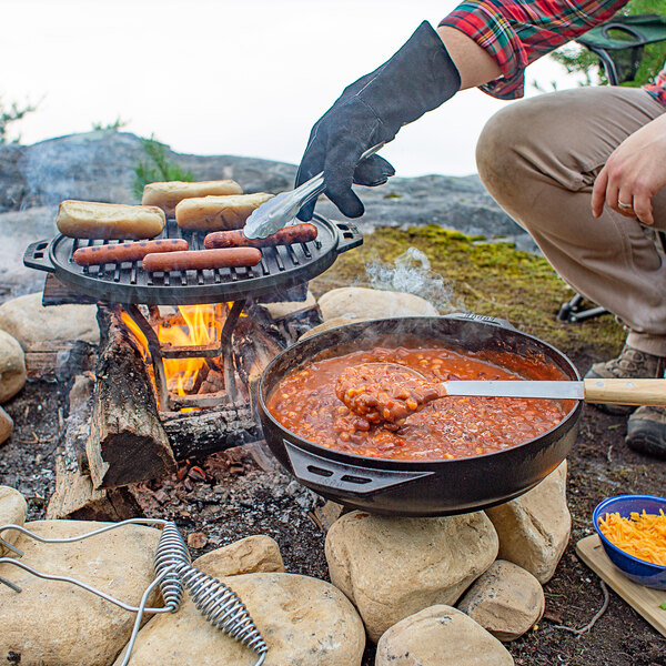 A person using a Lodge Cast Iron Cook-It-All to cook food over a campfire.