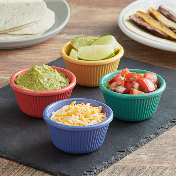 A group of Elite Global Solutions fluted melamine ramekins in assorted colors filled with salsa, guacamole, cheese, limes, and chopped tomatoes and onions.