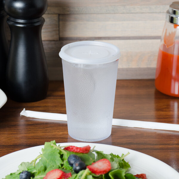 A Carlisle clear plastic tumbler filled with water on a table with a plate of fruit.