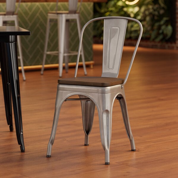 A Lancaster Table & Seating silver metal cafe chair with a walnut wood seat sits in front of a table.