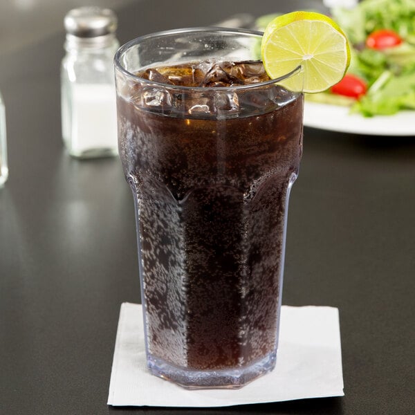 A Carlisle clear plastic tumbler filled with brown soda, ice, and a lime wedge on the rim.