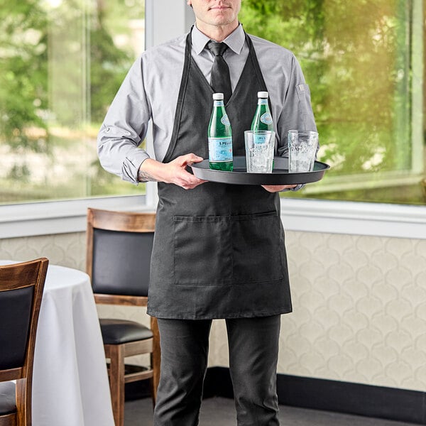 A man in a black Uncommon Chef tuxedo apron holding a tray with bottles and glasses.