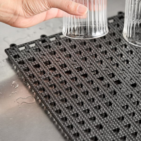 A hand pouring water into a glass over an American Metalcraft black vinyl interlocking bar tile.