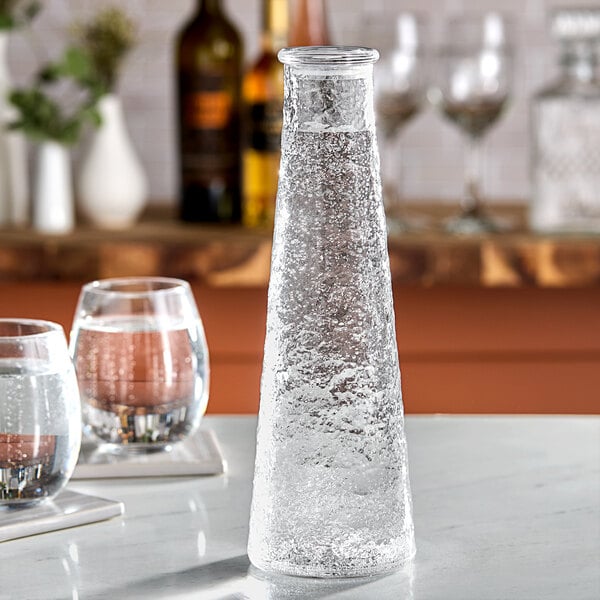 An American Metalcraft clear pebbled acrylic water bottle on a table with a glass of water.