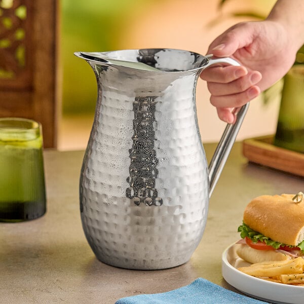 A hand holding a silver American Metalcraft bell pitcher with a handle.