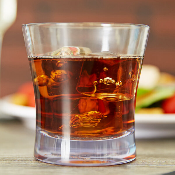 A Carlisle plastic rocks glass filled with brown liquid and ice on a table in a bar.