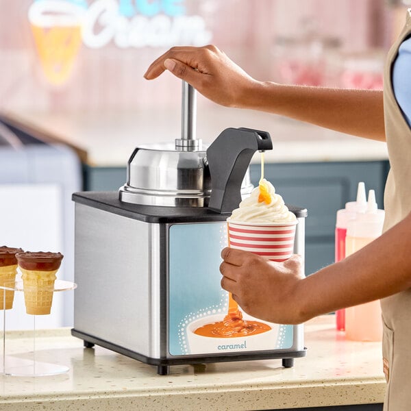 A stainless steel hot topping warmer with a pump dispenser being used to pour caramel sauce onto a cup of soft serve ice cream.
