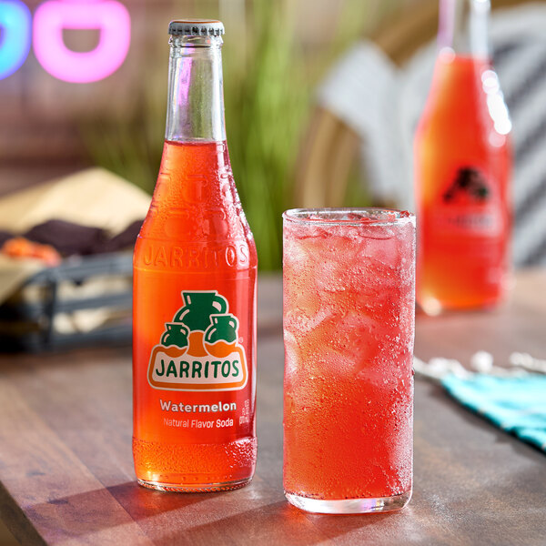 A bottle of Jarritos watermelon soda next to a glass filled with ice and the same red soda on a table.