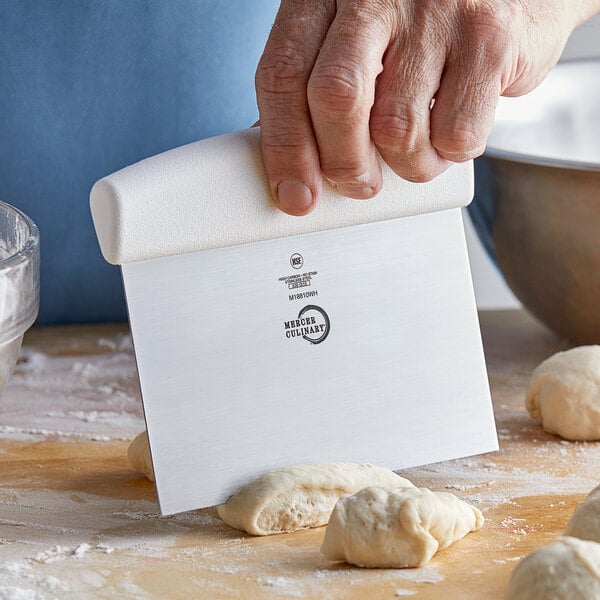 A person using a white-handled Mercer Culinary dough cutter to cut dough on a wooden table.
