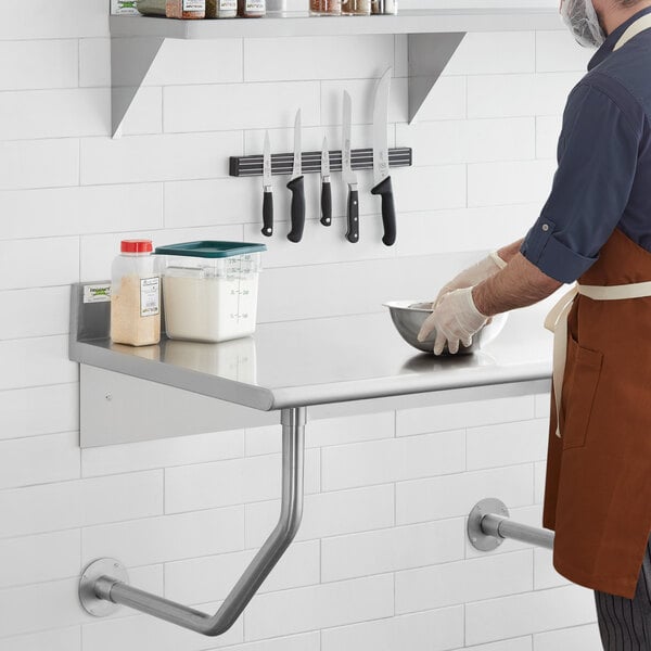 A man in a brown apron using a Regency stainless steel wall mounted table in a professional kitchen.