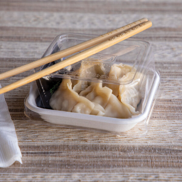 A clear, square compostable plastic lid covering a small takeout container of dumplings with chopsticks placed on top.