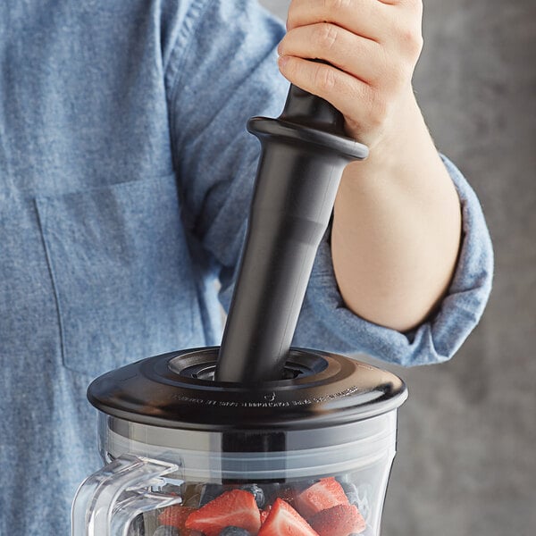 hand using a black tamper to push ingredients into a blender jar