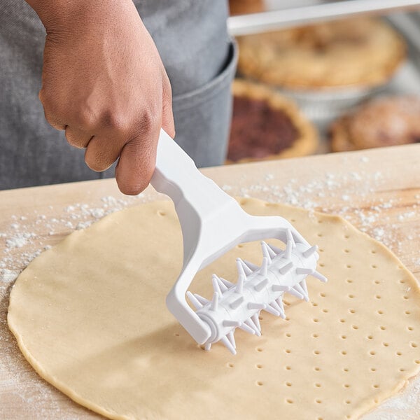 A white plastic dough docker with a handle and spiked rollers being used on rolled-out dough.