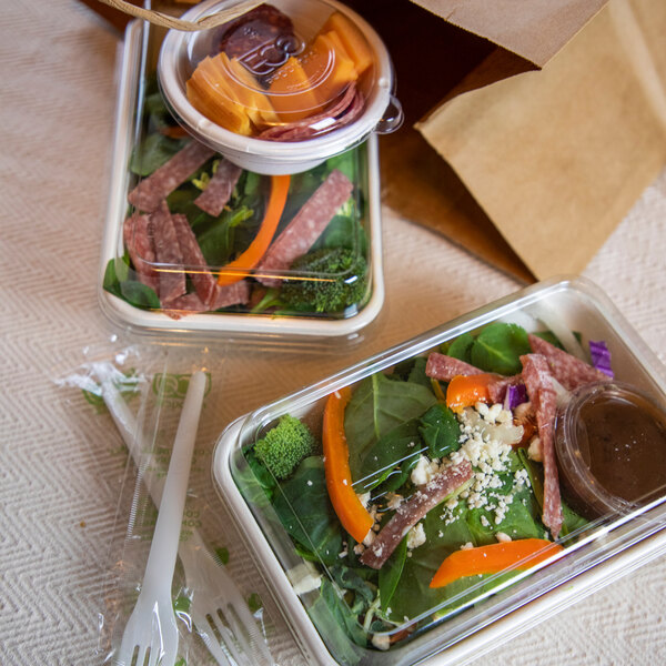 A set of clear rectangular plastic lids covering takeout salad containers, shown with utensils and a brown paper bag.