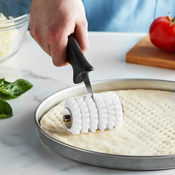 A hand holding an American Metalcraft dough docker with pointed wheels and a black plastic handle above pizza dough.