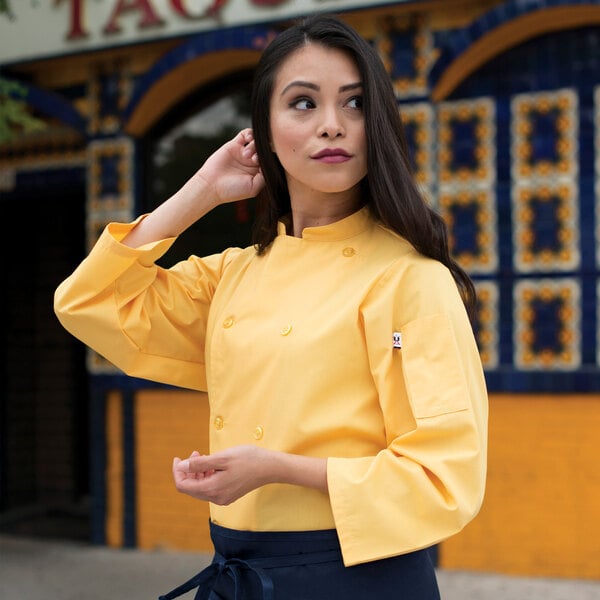 A woman in a yellow chef's jacket poses for a photo on a counter in a professional kitchen.