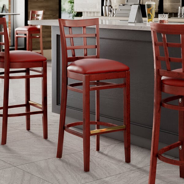 A group of Lancaster Table & Seating mahogany wood bar stools with burgundy vinyl seats next to a bar counter.