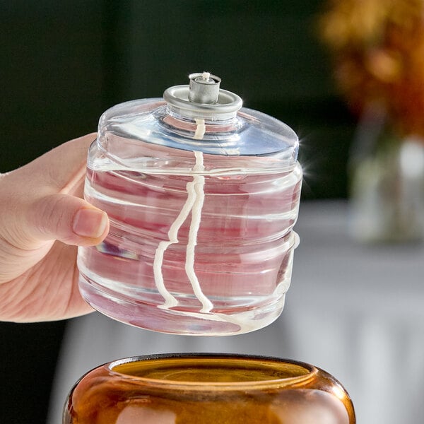 A clear liquid candle fuel cartridge with a visible wick and metal top being held above a container.