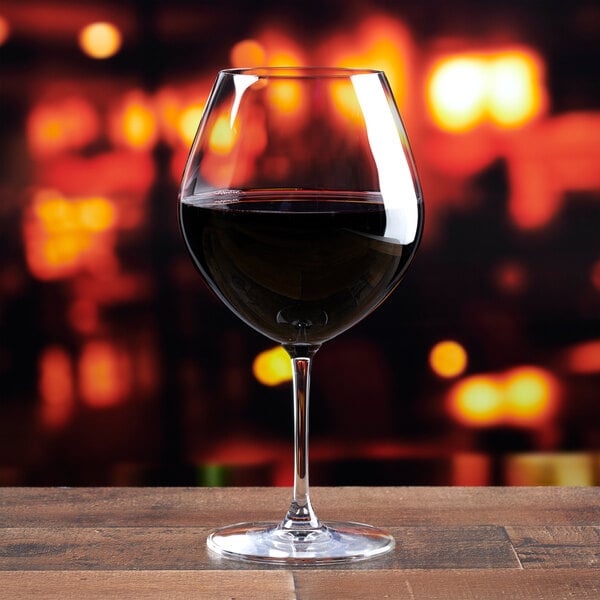A clear, stemmed Burgundy wine glass filled with red wine, placed on a wooden surface with a blurred warm-toned background.