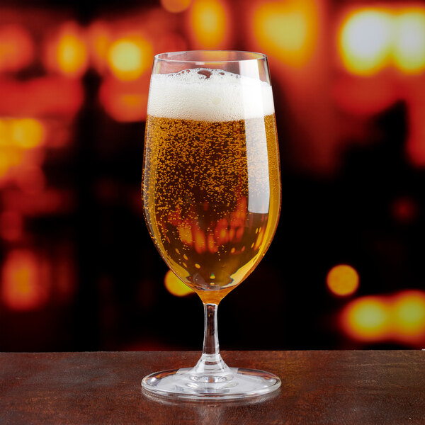 A clear 13.6 oz. water goblet filled with a carbonated beverage, set against a blurred warm-toned background.