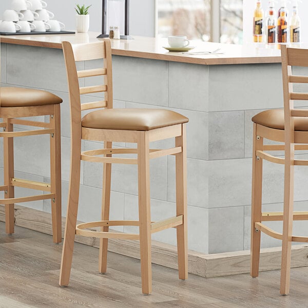 Three Lancaster Table & Seating natural wood ladder back bar stools with light brown vinyl seats at a counter.