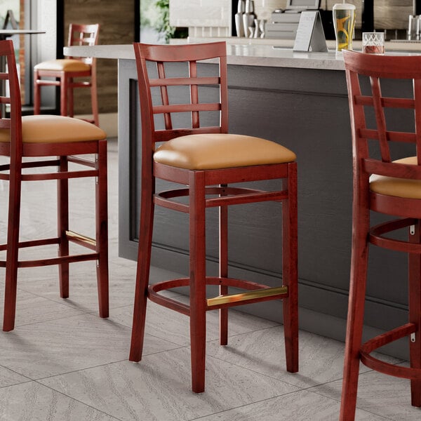 A group of Lancaster Table & Seating mahogany wood window back bar stools with light brown vinyl seats next to a counter.