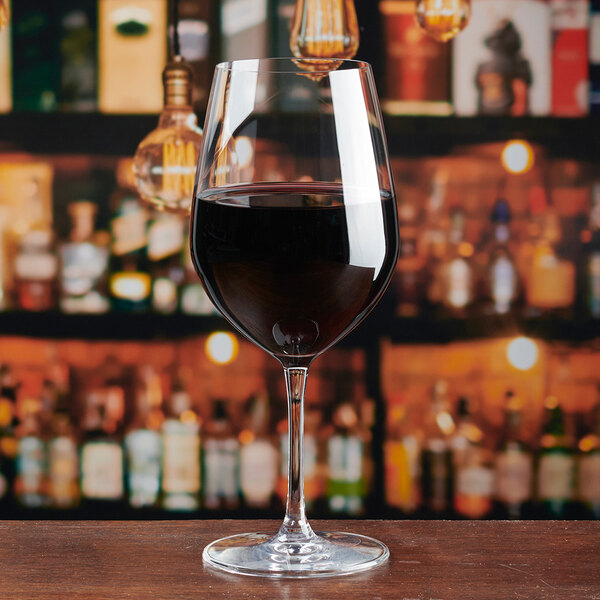 A clear Lucaris Temptation 20.5 oz. Bordeaux wine glass filled with red wine, set on a wooden surface with a blurred bar background.