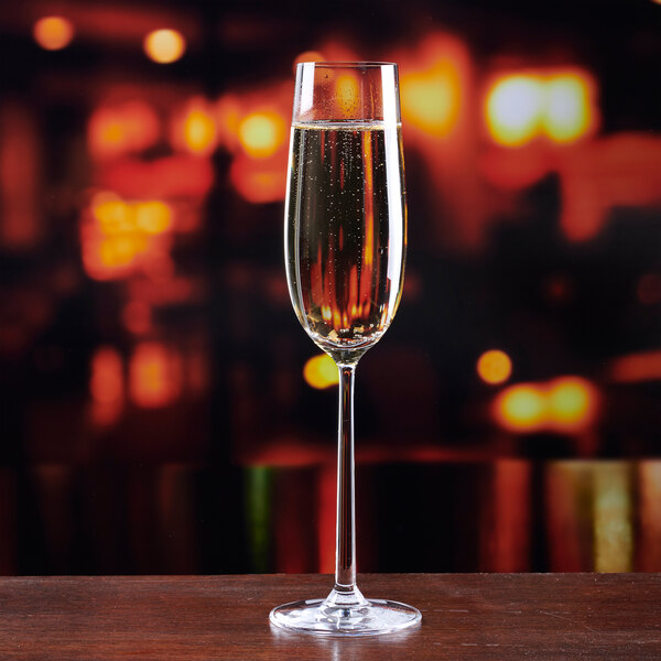 A clear 8.5 oz. champagne flute filled with sparkling wine, standing on a wooden surface with a blurred warm-toned background.