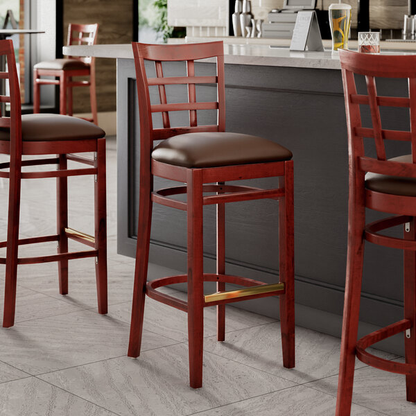 Three Lancaster Table & Seating mahogany wood bar stools with dark brown vinyl seats next to a counter.