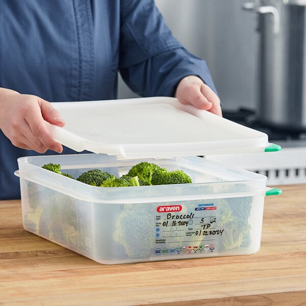 A woman holding a Araven translucent plastic food pan with broccoli in it and the lid open.