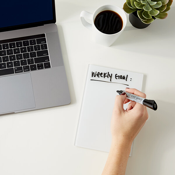 A hand using a white marker to write on a white Quartet glass notepad on a desktop with a laptop.