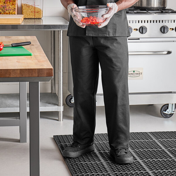 A person wearing Uncommon Chef black classic pants in a professional kitchen holds a bowl of food.