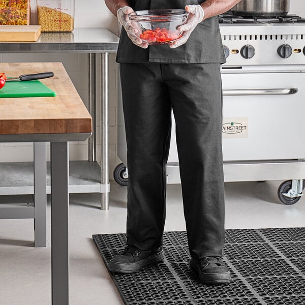 A person wearing Uncommon Chef black pants in a professional kitchen holding a bowl of food.