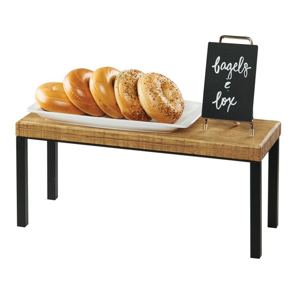 A rectangular rustic pine wood riser with black metal legs, measuring 20 inches by 7 inches by 9 inches, shown holding a plate of bagels and a small sign.