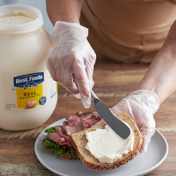 A large 1-gallon container of Best Foods Real Mayonnaise next to a person spreading mayonnaise on a sandwich.