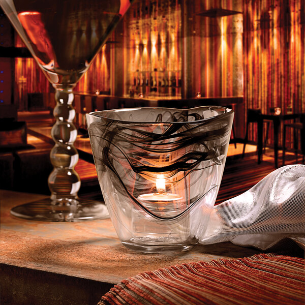 A glass with a Hollowick Black Glass Votive with a candle in it on a table in a room with a window.