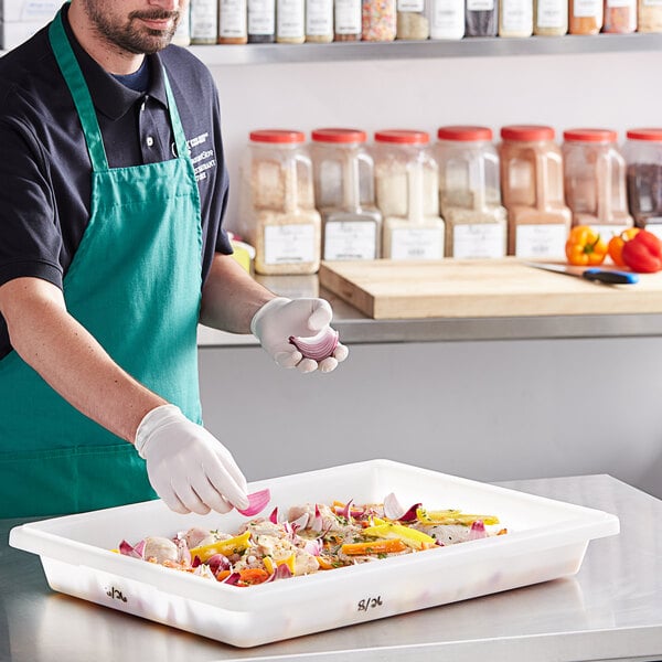 A man in a green apron and gloves using a white Vigor polyethylene food storage container to hold food.