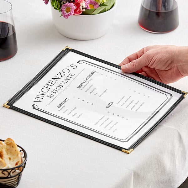 A hand holding a H. Risch, Inc. black vinyl menu cover with gold corners on a table.