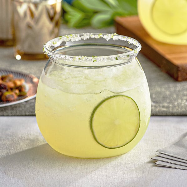 A clear plastic stemless fish bowl glass filled with a yellow beverage, garnished with a lime slice and a salted rim.