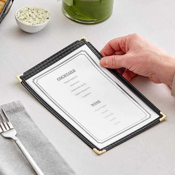 A hand holding a black H. Risch, Inc. vinyl menu cover with gold corners on a table.