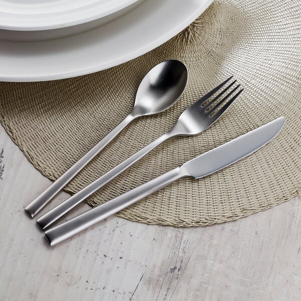 A Sola stainless steel serving spoon on a placemat with a fork and knife.