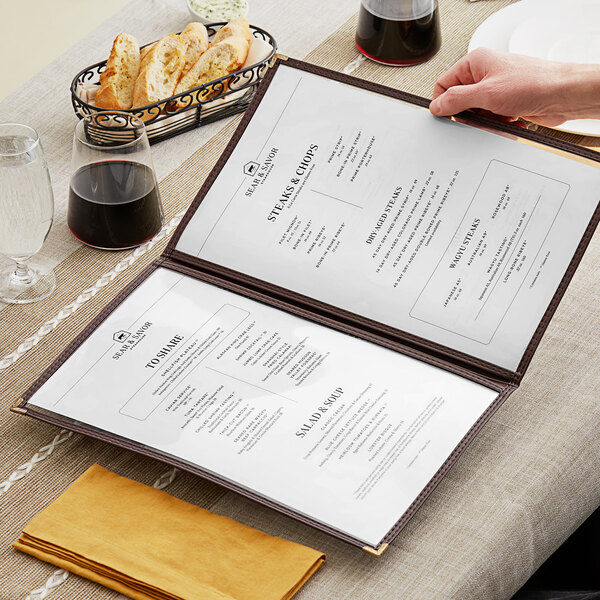 A hand holding a brown menu with gold corners on a table.