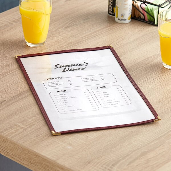 A maroon H. Risch, Inc. TES Deluxe vinyl menu cover on a table with a glass of orange juice and coffee.