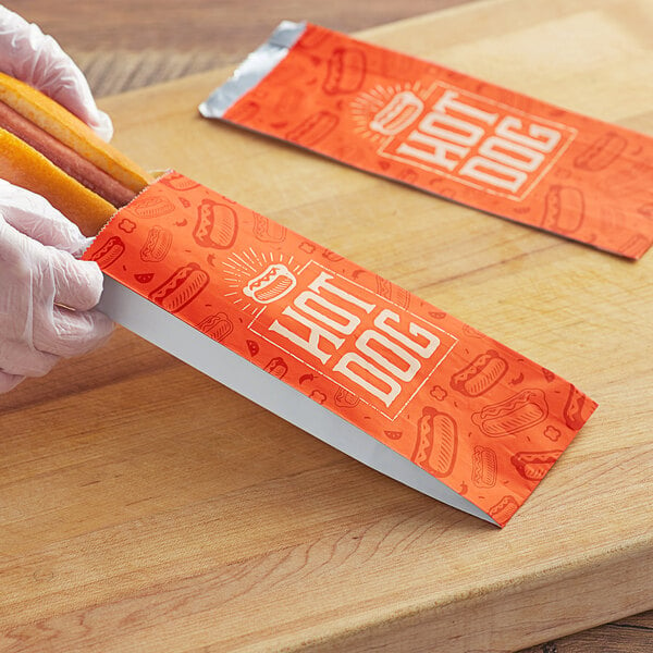 A person placing a hot dog in a red and white logoed paper bag.