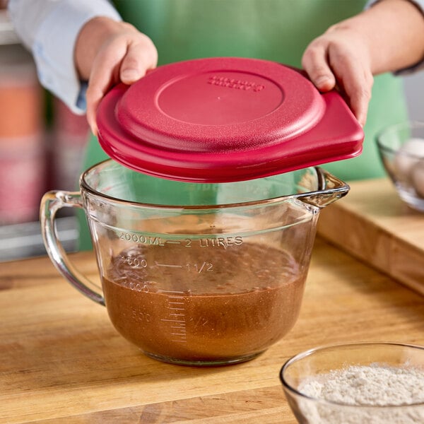 A person pouring brown liquid from a measuring cup with a red lid into a glass container.