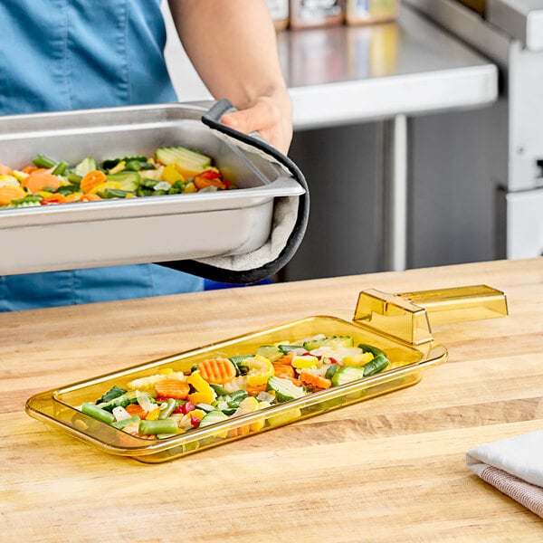 A woman using a Carlisle amber plastic food pan with handle to serve vegetables on a salad bar counter.