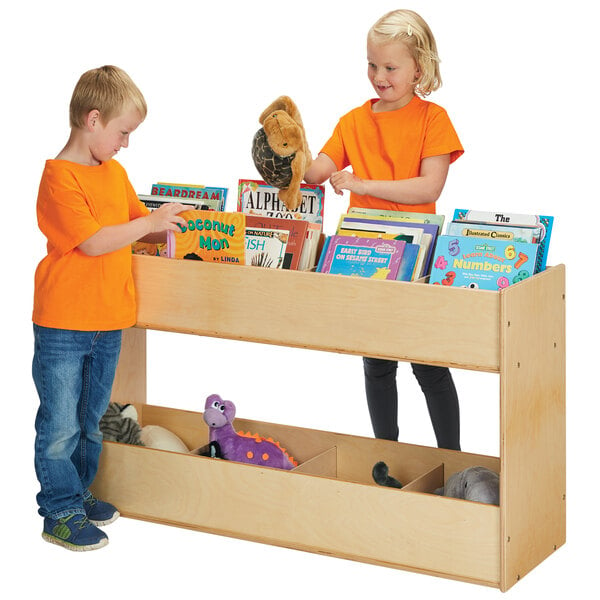 A boy and girl in orange and blue shirts standing next to a Young Time natural wooden bookshelf filled with books.